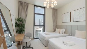 A modern hotel room with two beds, a desk, a chair, a large window with a city view, and a chandelier.