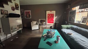 Une pièce avec une table de couleur turquoise, un lit, un fauteuil blanc et une affiche accrochée au mur.