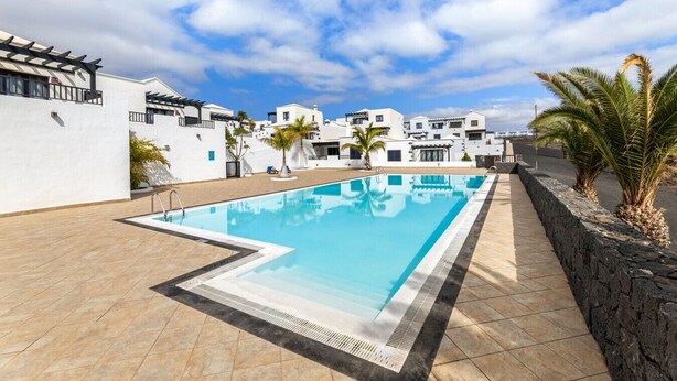 A swimming pool with a tiled deck, surrounded by white buildings with balconies, under a partly cloudy sky. - Casa Variza, tu refugio en Playa Blanca (Playa Blanca)