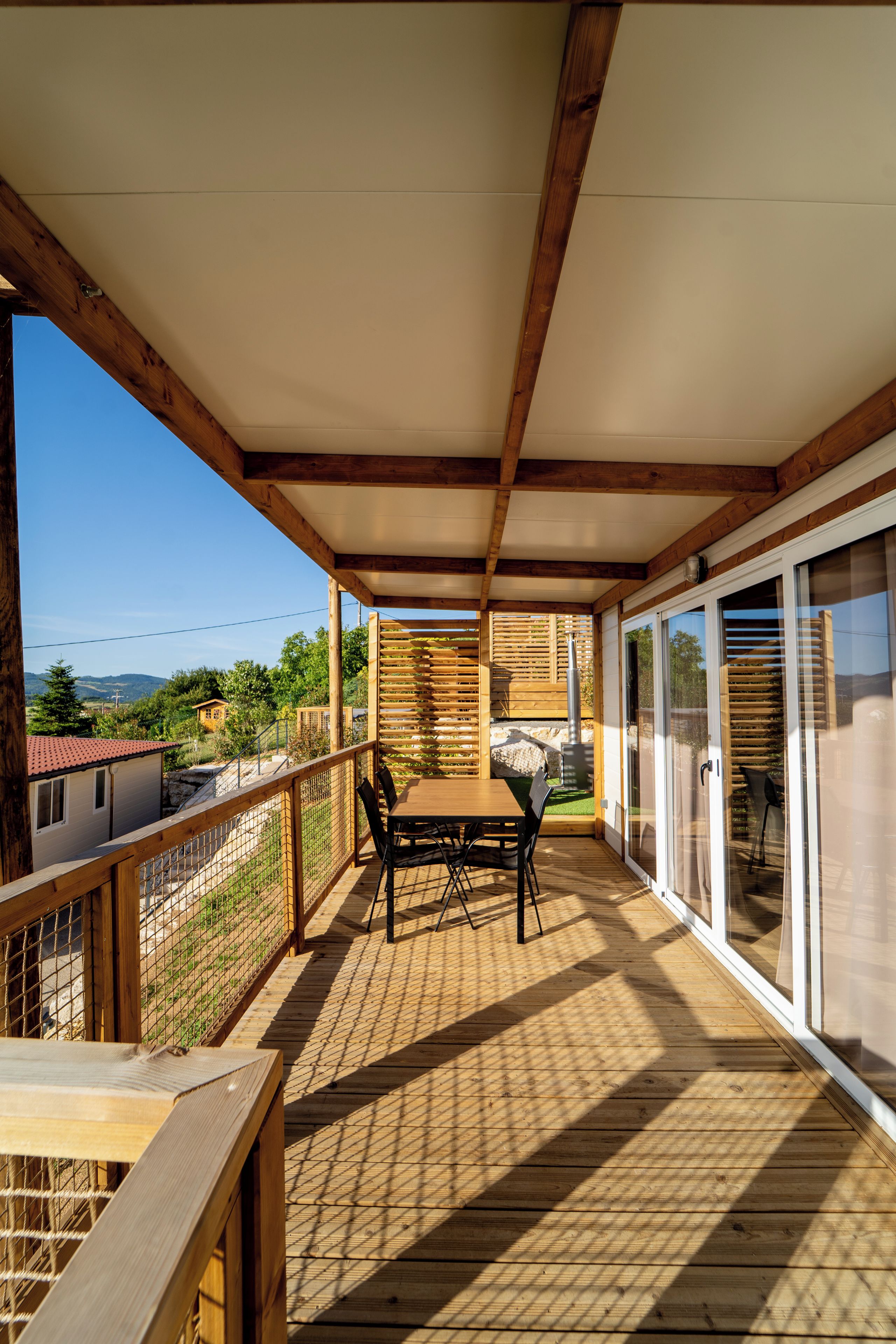 Una terraza de madera con una mesa y sillas, un vidrio que da al exterior y vistas del paisaje circundante.