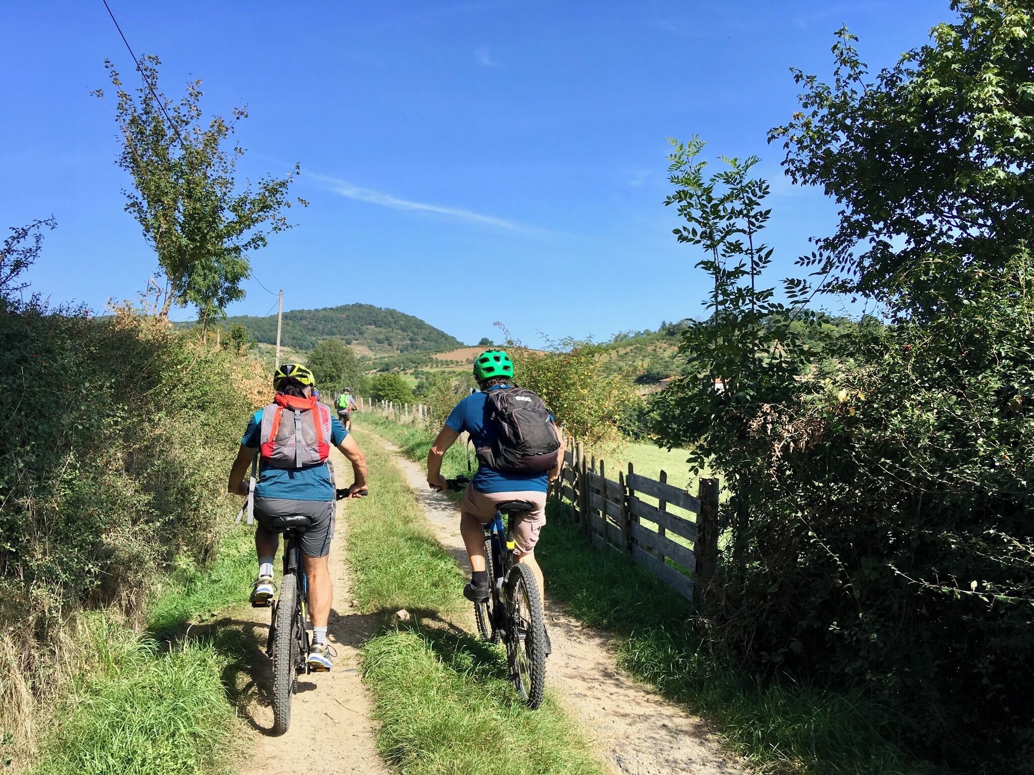Two cyclists on a dirt path surrounded by greenery and a wooden fence.
