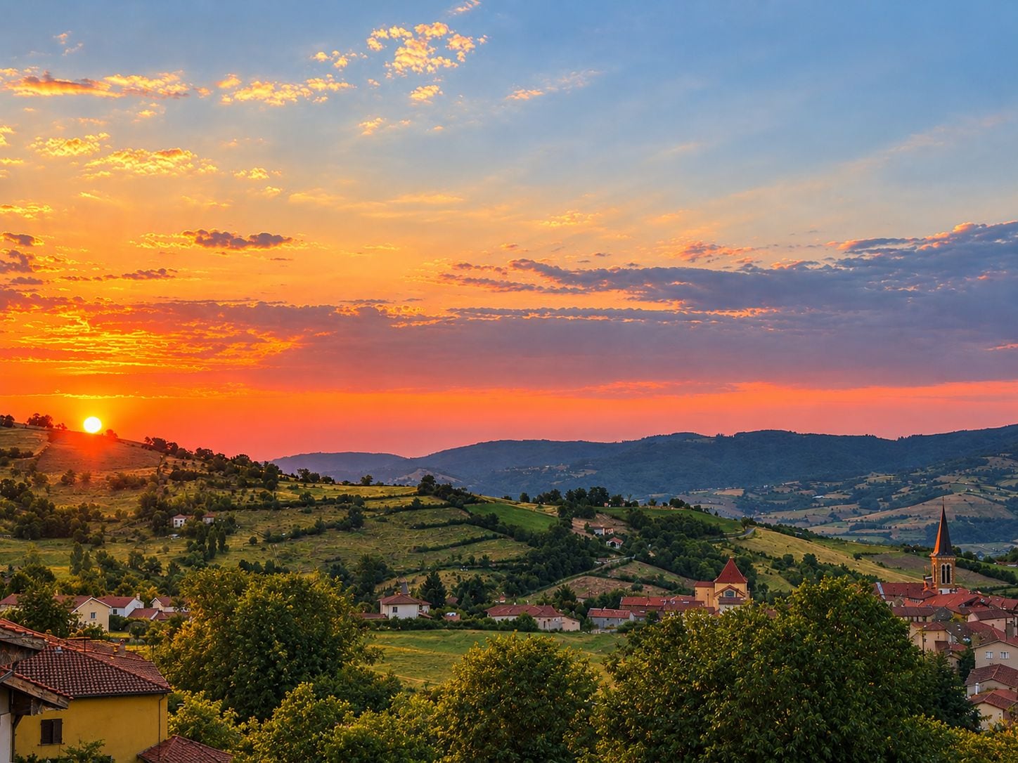 A rural landscape with rolling hills, a church, and a village during sunset.