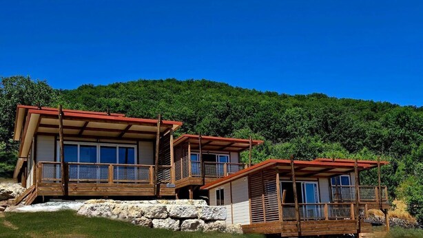 A two-story wooden cabin with a red roof, situated on a rocky hillside with a dense forest backdrop. - Tiny d'Monts (Brullioles)