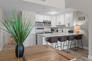 A modern kitchen with white cabinets, a stainless steel refrigerator, a microwave, and a coffee maker. There is a wooden dining table with a vase of green plants.