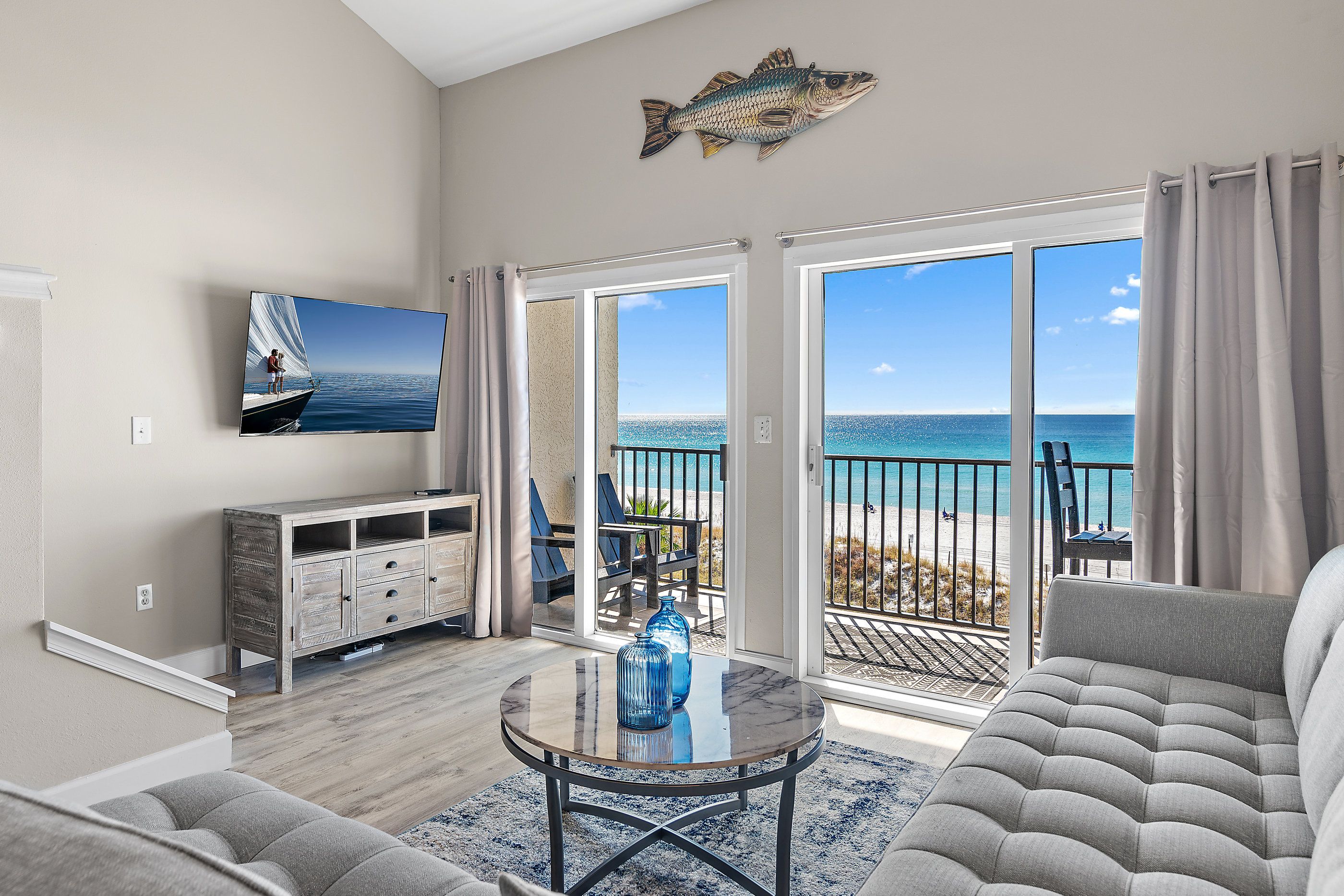 A living room with a sofa, a coffee table, and a TV on the wall. There is a balcony with a view of the ocean.