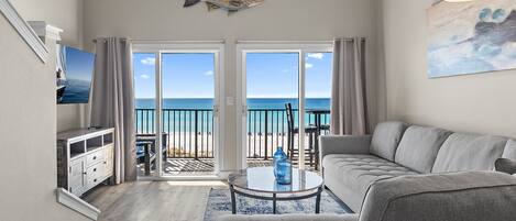 A living room with a sofa, a coffee table, and a TV. There is a balcony with a view of the ocean.