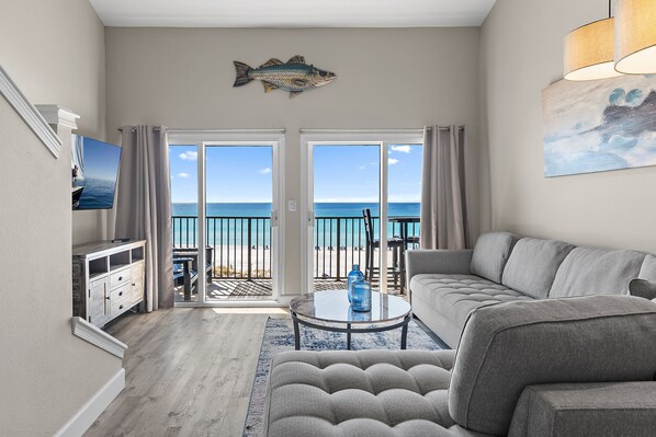 A living room with a sofa, a coffee table, and a TV. There is a balcony with a view of the ocean.