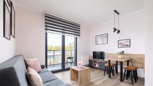 Ein modernes Wohnzimmer mit einem grauen Sofa, einem Holztisch und einem Fernseher auf einem Holzständer. Zwei Holzschemel und ein großes Fenster mit Jalousien sind ebenfalls vorhanden.