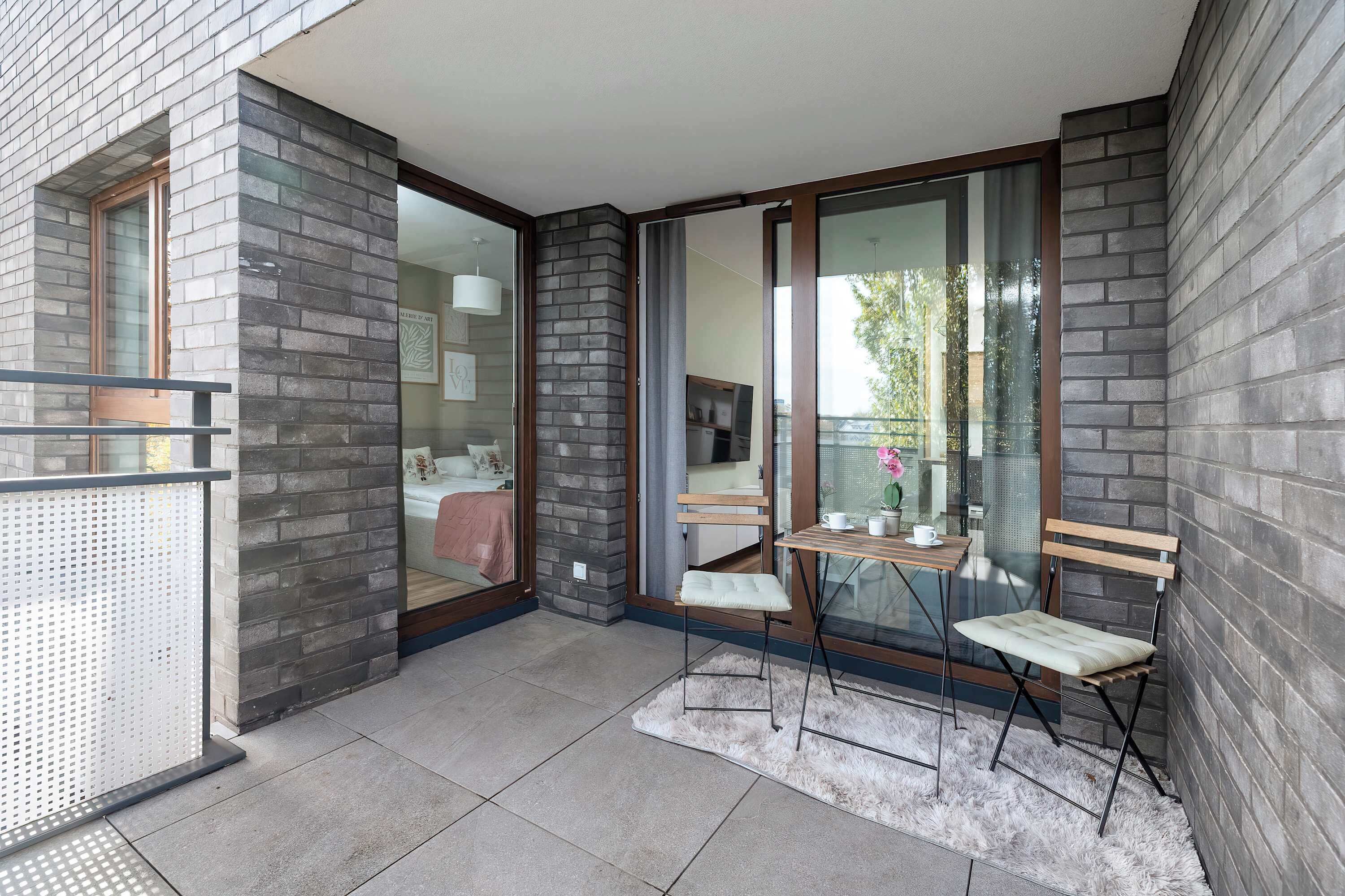 Un balcon moderne avec des murs en briques grises, une porte vitrée donnant sur une chambre à coucher et une petite table avec une chaise.