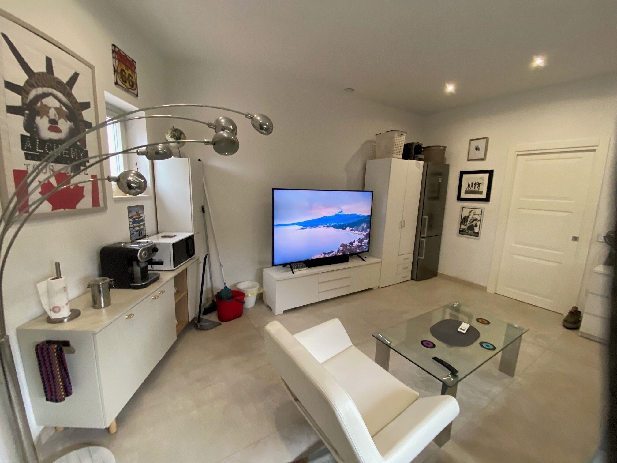 A modern living room with a flat-screen TV, white furniture, a glass coffee table, and a mounted lamp.