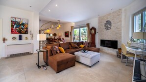 A modern living room with a sectional sofa, a coffee table, and a fireplace. There is a dining area with a table and chairs, and a kitchen visible in the background. - Pink Moon Villa. Private Pool. Close to the Beach. 10 guests. Luxury. (Fontane Bianche)