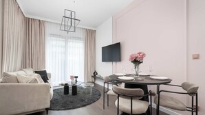A modern living room with a sofa, a dining table with chairs, a TV mounted on the wall, and a window with curtains.