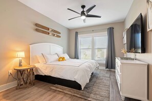 Une chambre à coucher comprenant un lit, une table de chevet, une lampe, un ventilateur de plafond, une fenêtre avec des rideaux et une commode.