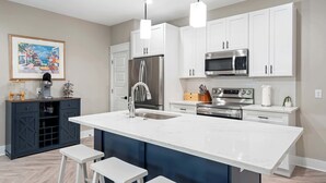 Une cuisine moderne avec des meubles blancs, un grand îlot central doté de tabourets, des appareils en acier inoxydable et un tableau accroché au mur.