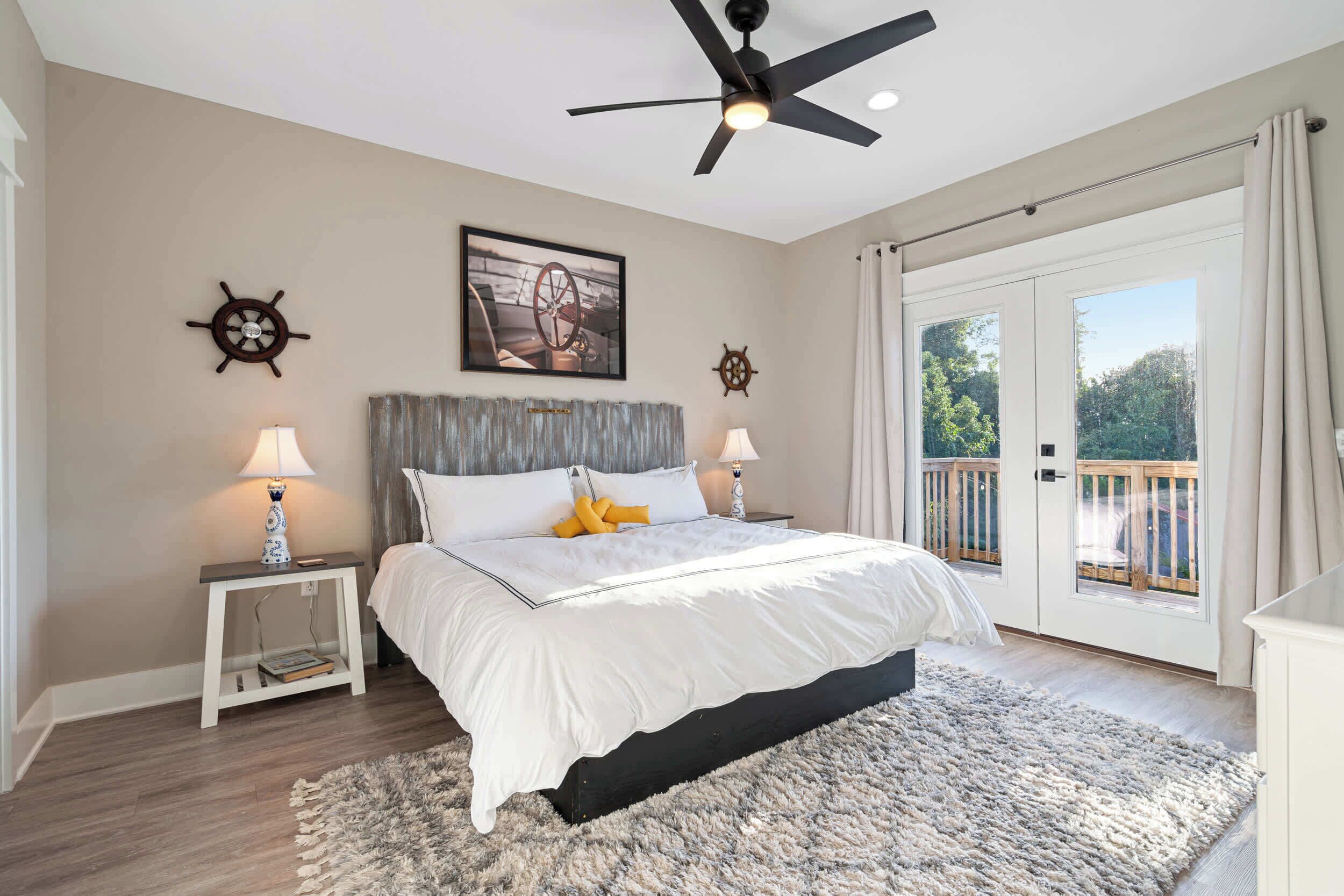 A bedroom with a bed, two bedside tables, a ceiling fan, a large window with a view, and a door leading to an outdoor area.