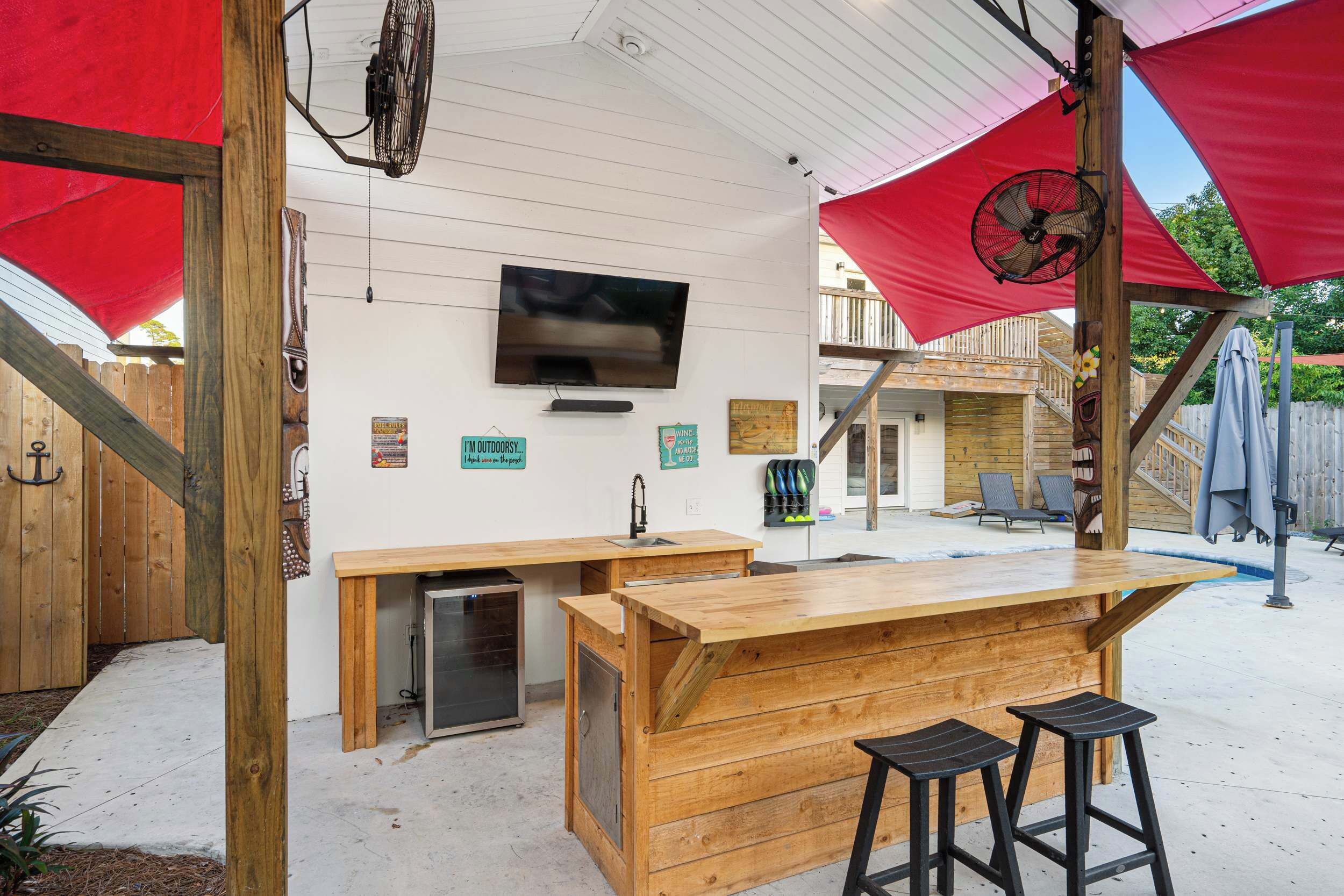 Un espace bar extérieur doté d’un comptoir en bois, de deux tabourets, d’un téléviseur fixé au mur et d’un parasol rouge offrant de l’ombre.