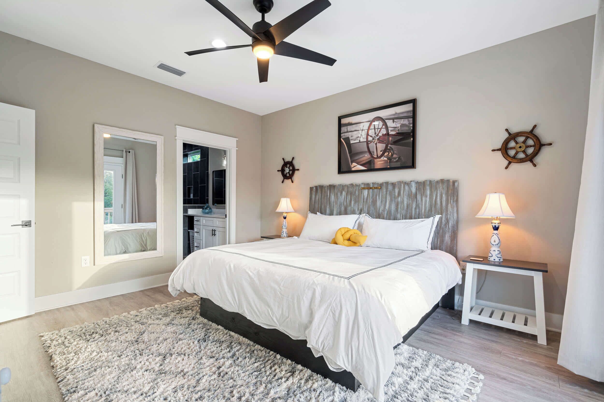 A bedroom with a bed, two bedside tables, a ceiling fan, a ship's wheel decoration, and a framed picture on the wall.