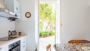 A kitchen with a built-in oven, a wooden table with a patterned tablecloth, and a view of a garden through an open door. - Spacious 2-bedroom flat near the beach (Rapallo)