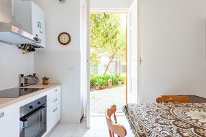 A kitchen with a built-in oven, a wooden table with a patterned tablecloth, and a view of a garden through an open door.