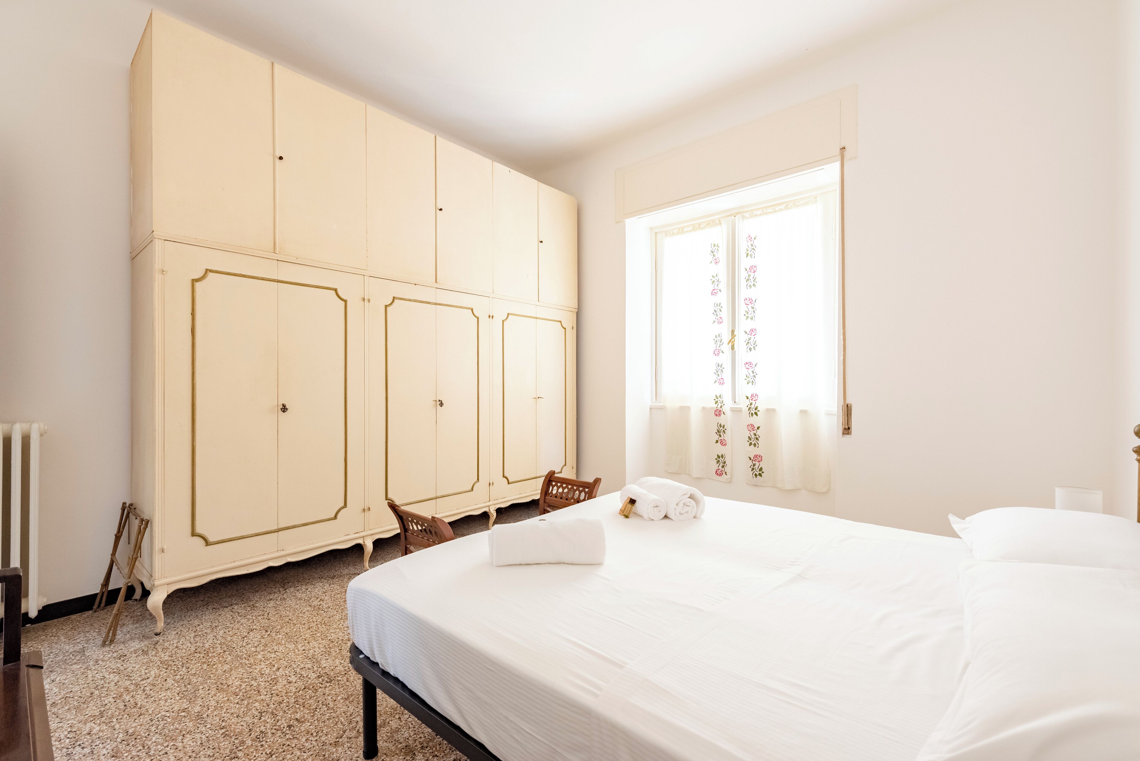 A bedroom with a large bed, a wardrobe, a chair, and a window with curtains.