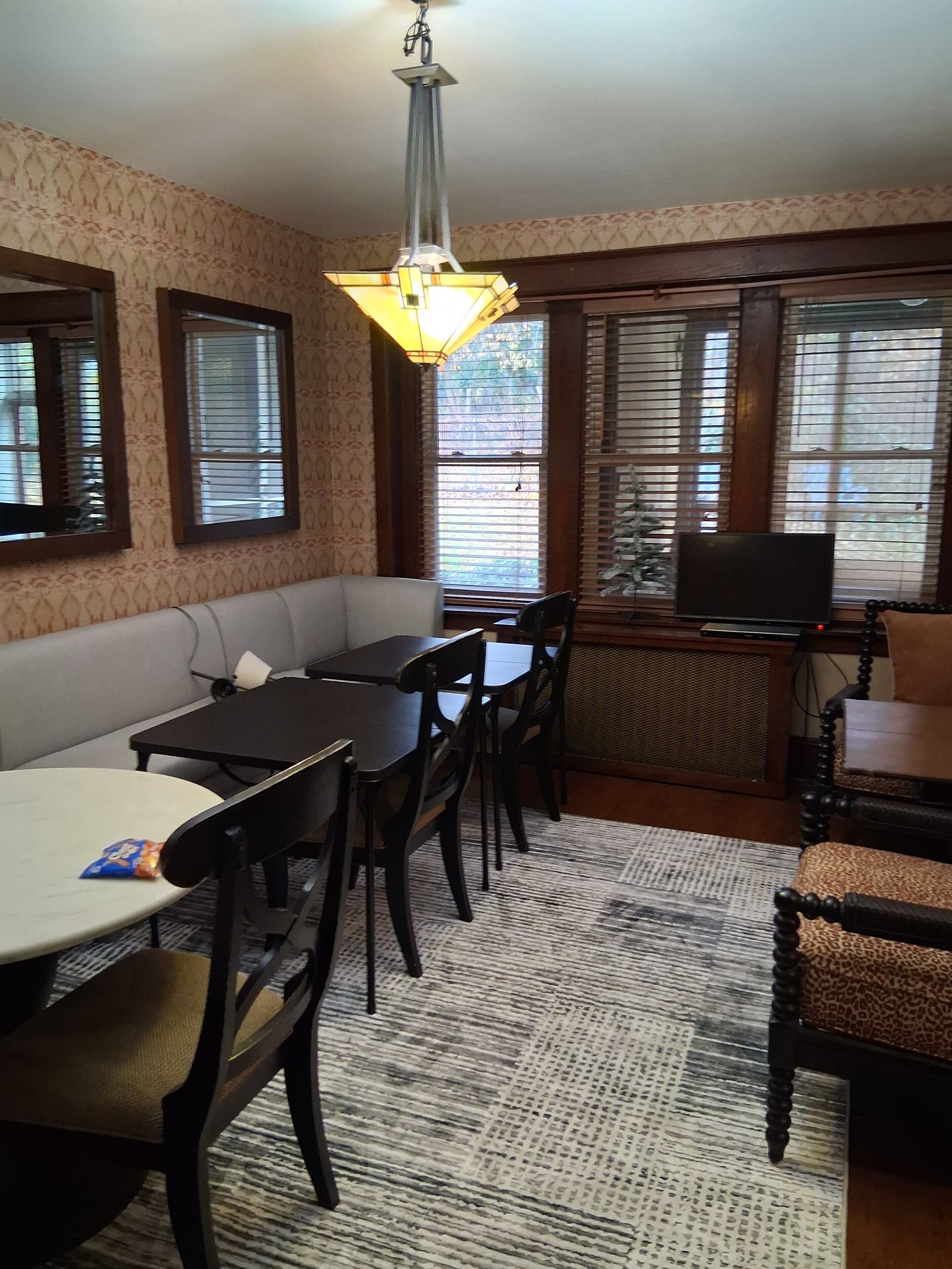 A room with a dining table, chairs, a sofa, a television, and windows with blinds.