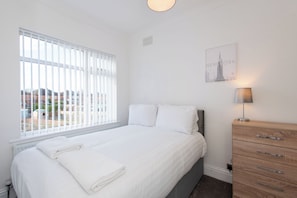 Une chambre avec une grande fenêtre, un lit recouvert de draps blancs, une commode en bois et une lampe fixée au mur.