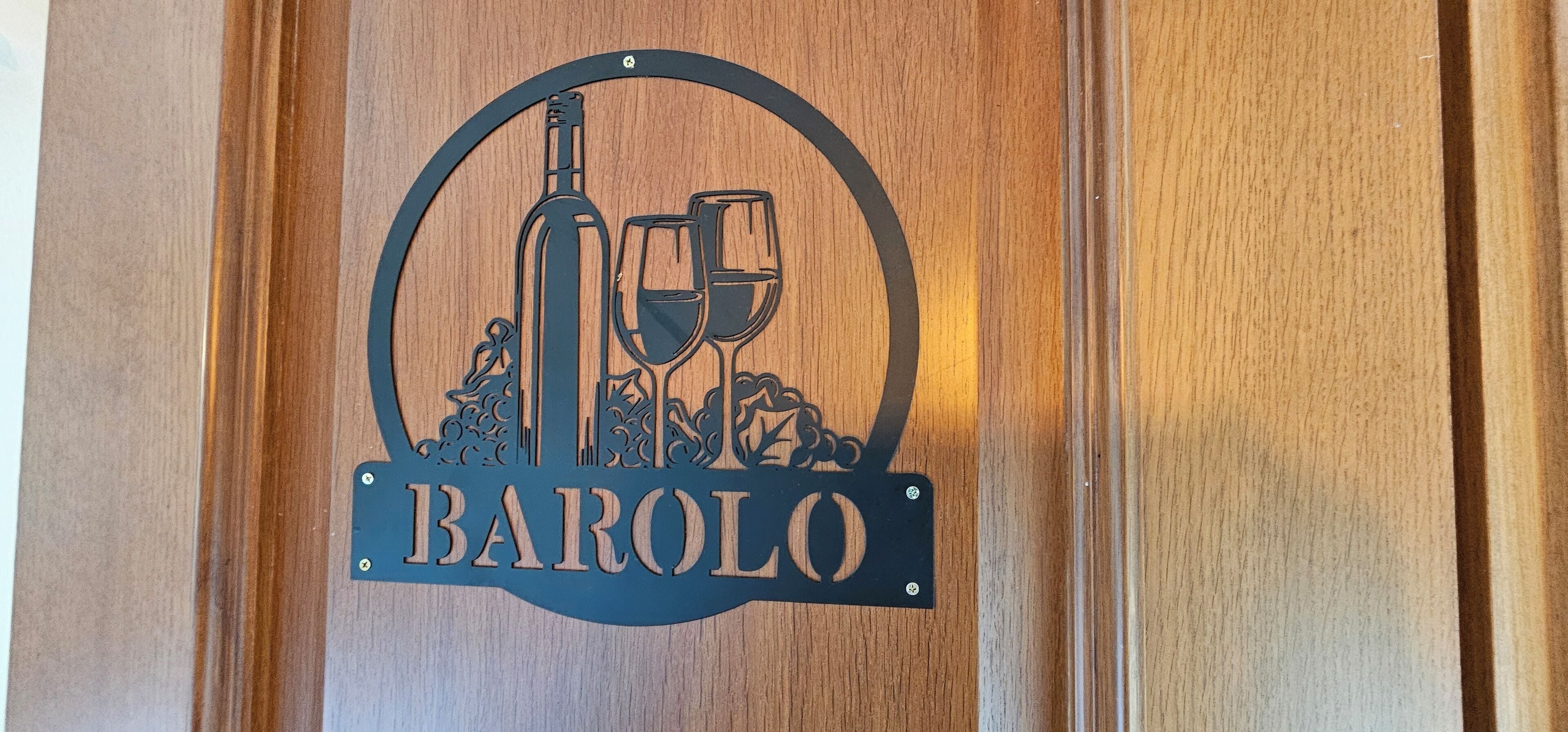 Un letrero de madera con un logotipo circular que muestra una botella de vino y copas, y la palabra 'BAROLO' debajo.