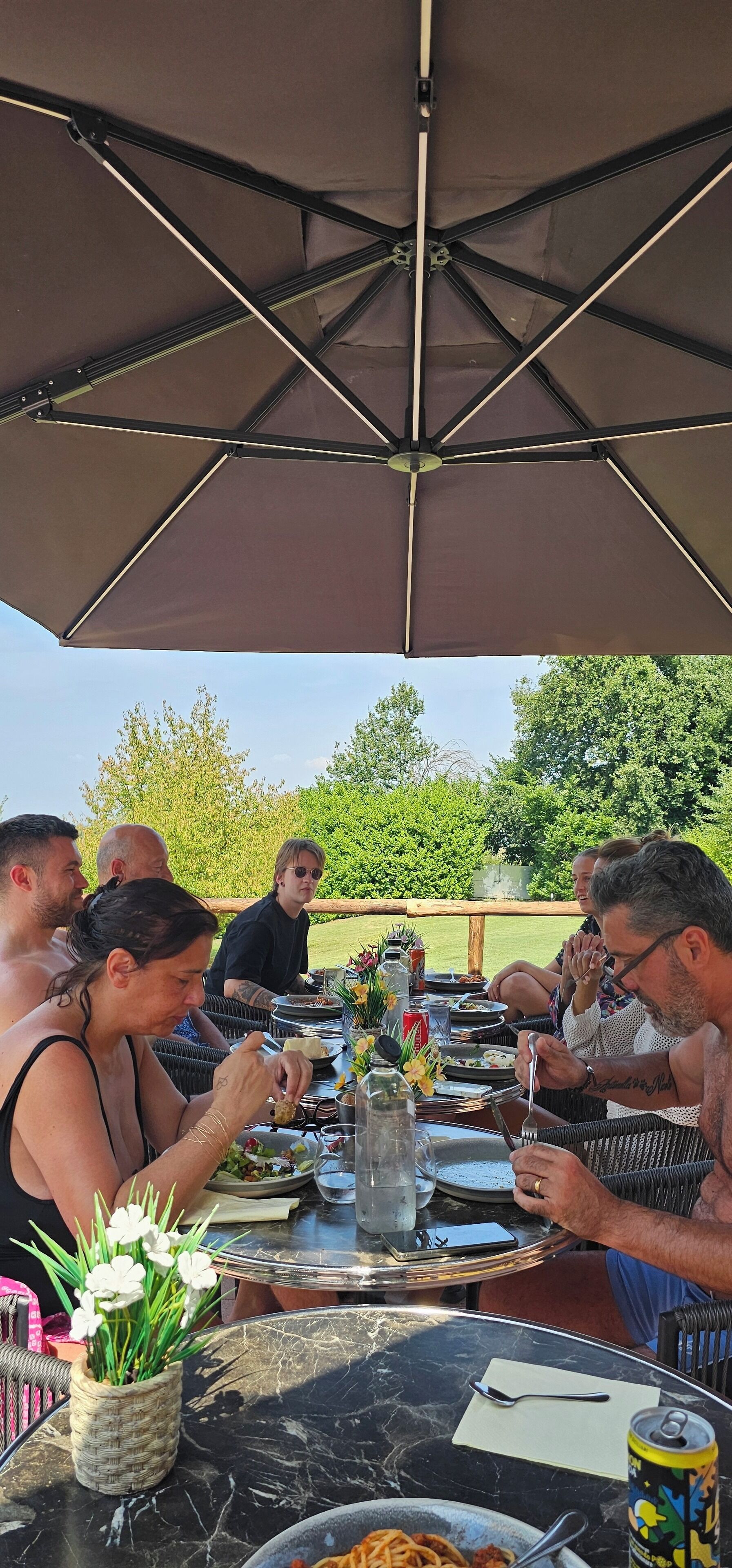 Un grupo de personas cenando en una mesa al aire libre, bajo un gran paraguas, rodeado de vegetación.