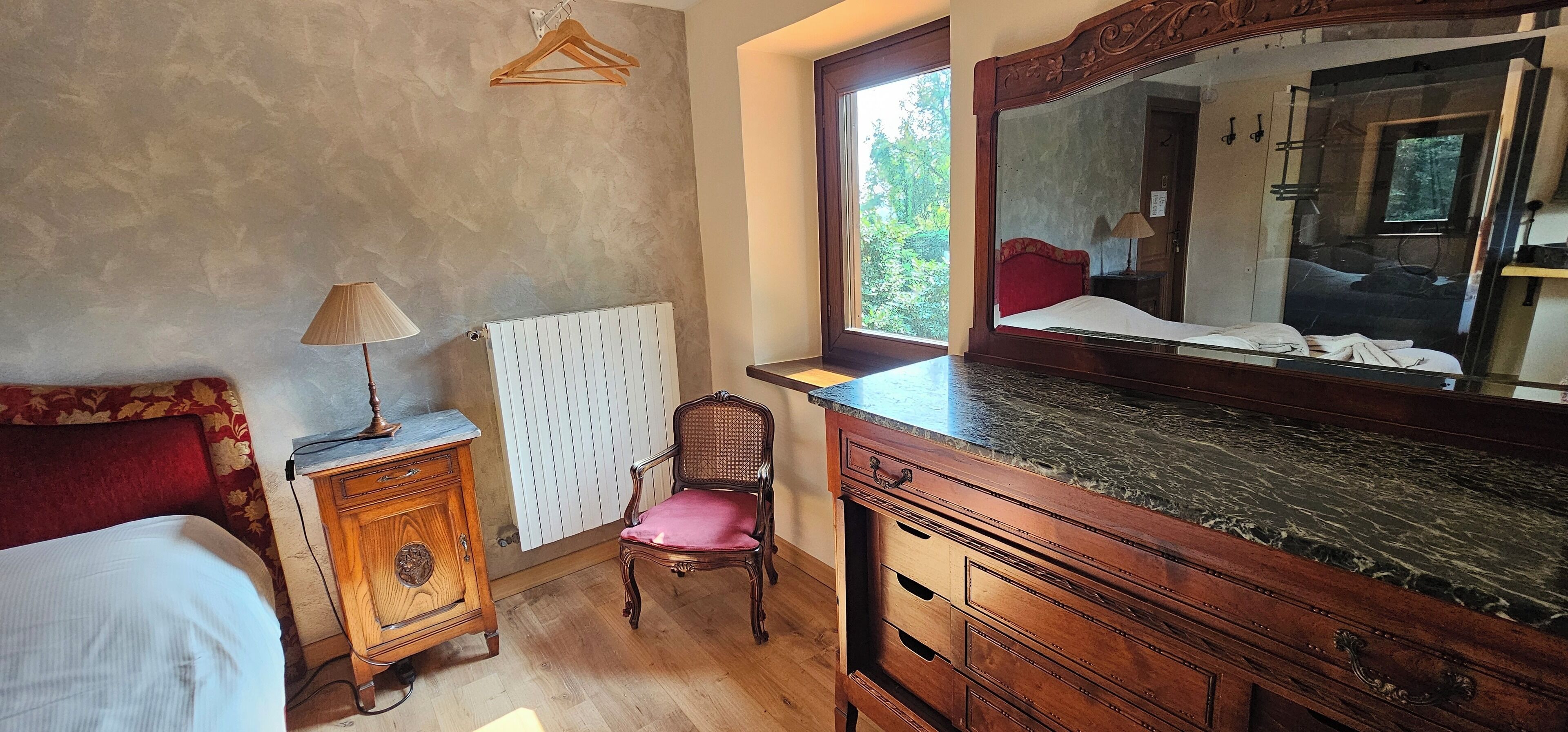 Une chambre à coucher avec un lit, une commode en bois, une chaise, un radiateur et un grand miroir.