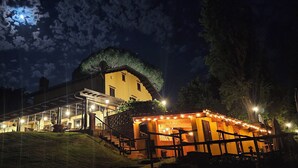 A large, well-lit building with a prominent tree on the roof, a covered outdoor area with string lights, and a staircase leading to the entrance. - Villa Fonte  (Ferrere)