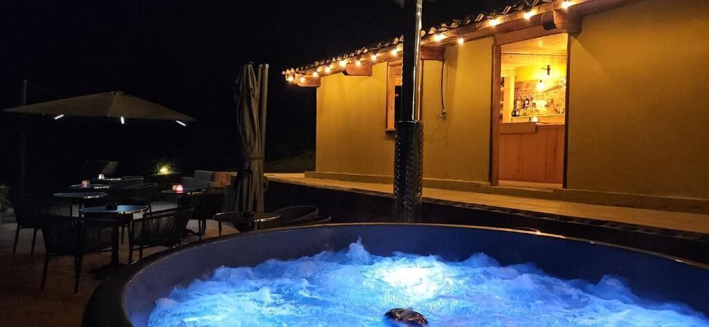 A hot tub with a person relaxing, outdoor dining area with tables and chairs, and a building with a bar visible through an open door.