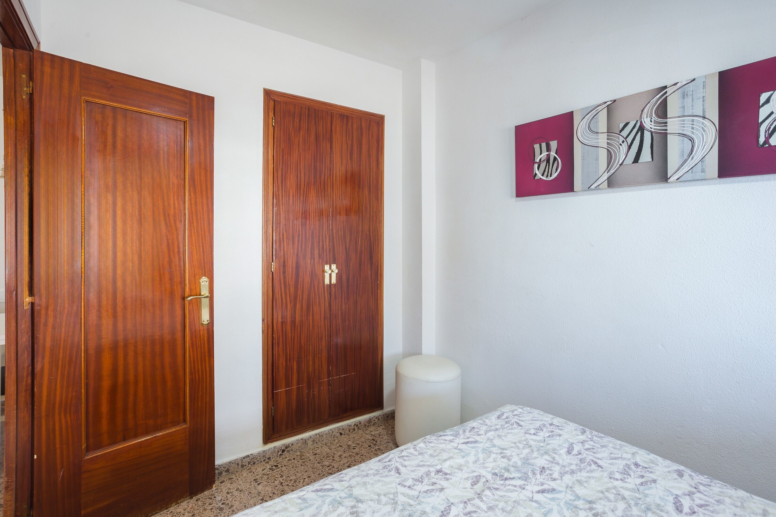 A room with two wooden doors, a white wall with abstract art, a patterned bedspread, and a small white trash bin.