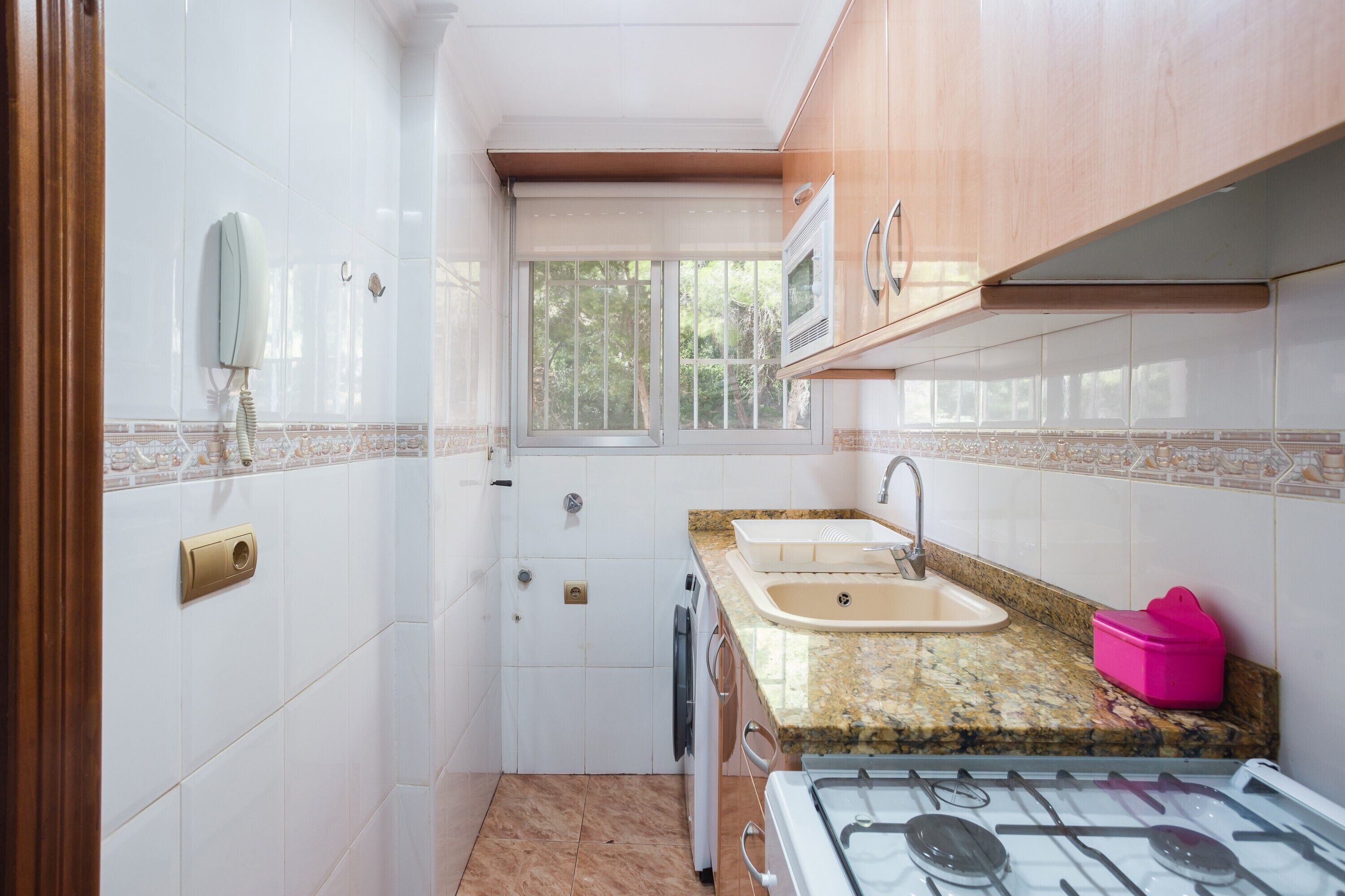 Una cocina compacta con azulejos blancos, estufa a gas, microondas, pileta y ventana.