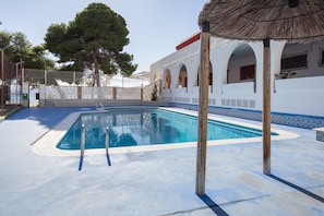 A swimming pool with a tiled deck, a wooden pergola, and a white building with arches.