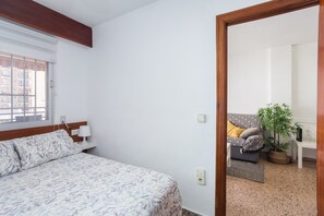 Ein Schlafzimmer mit Bett, Nachttisch, Lampe, Fenster und Blick auf einen Wohnbereich mit Sofa und Pflanze.