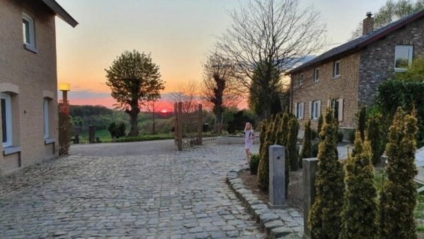 Ein Kopfsteinpflasterweg führt zu einem Gebäude mit einer Stein-Fassade, umgeben von Bäumen und einem Sonnenuntergang im Hintergrund.