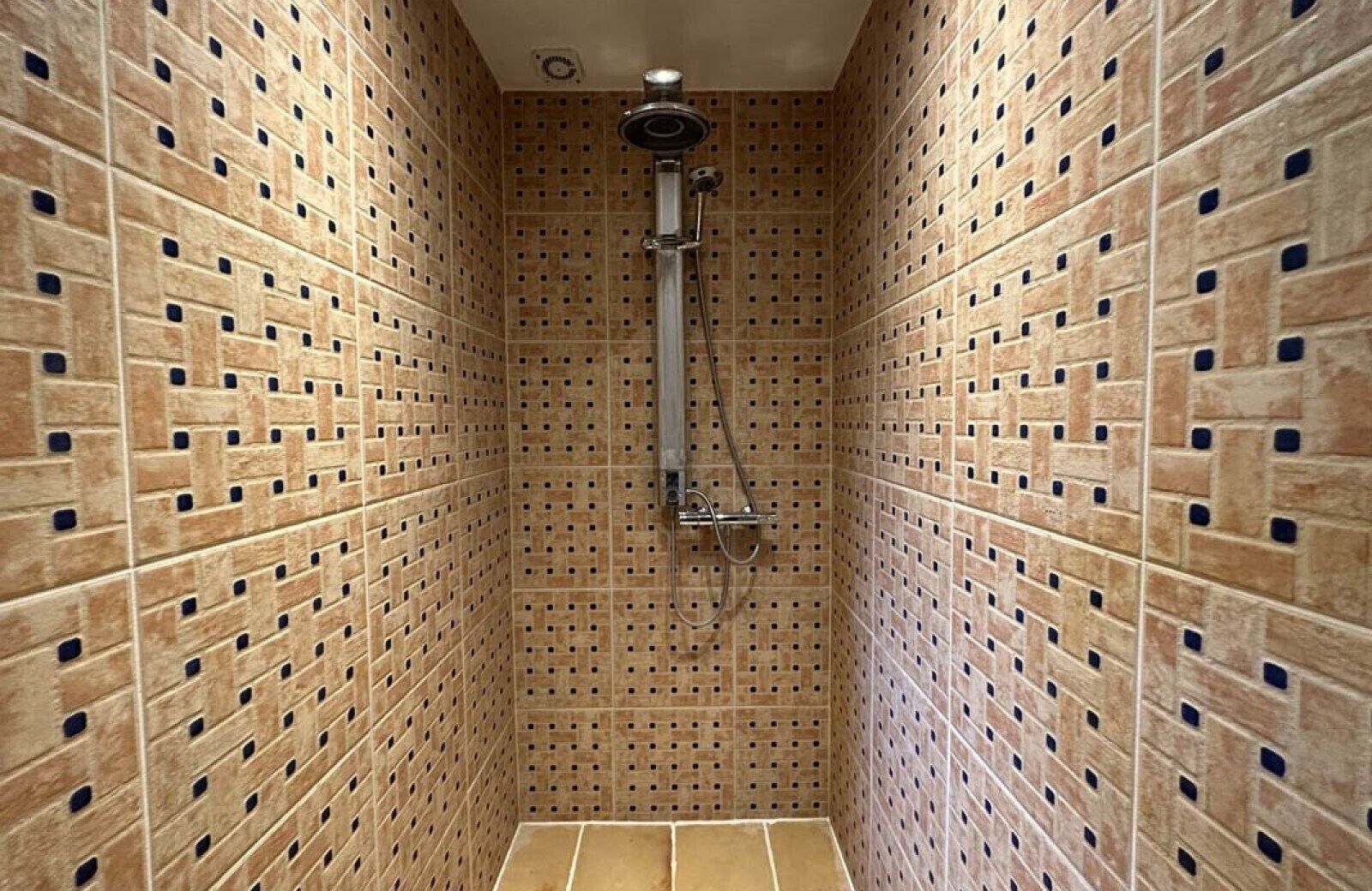 A narrow shower stall with a tiled wall and floor, a showerhead, and a handheld shower attachment.