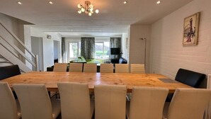 A dining area with a large wooden table and beige chairs, a staircase on the left, and a kitchen in the background.