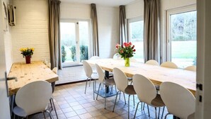 A dining area with a wooden table and white chairs, a vase with flowers, and a view of greenery outside.