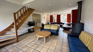 A spacious living area with a wooden staircase, a sofa set, a round wooden table, and a black wood-burning stove.