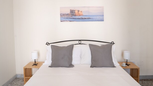 A neatly made bed with two pillows, a picture of a coastal building on the wall, and two bedside tables with lamps.