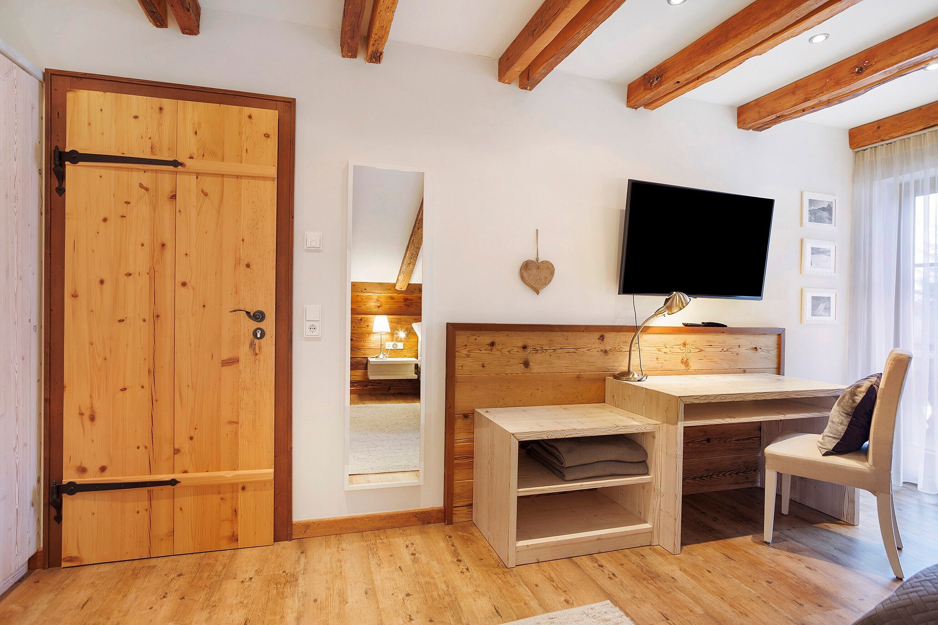 A room with a wooden door, a bed with a headboard, a desk with a chair, a television mounted on the wall, and a window with curtains.
