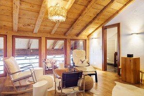 Un interior de madera con una mecedora, una silla blanca con un cojín de ciervo y una mesa de madera.