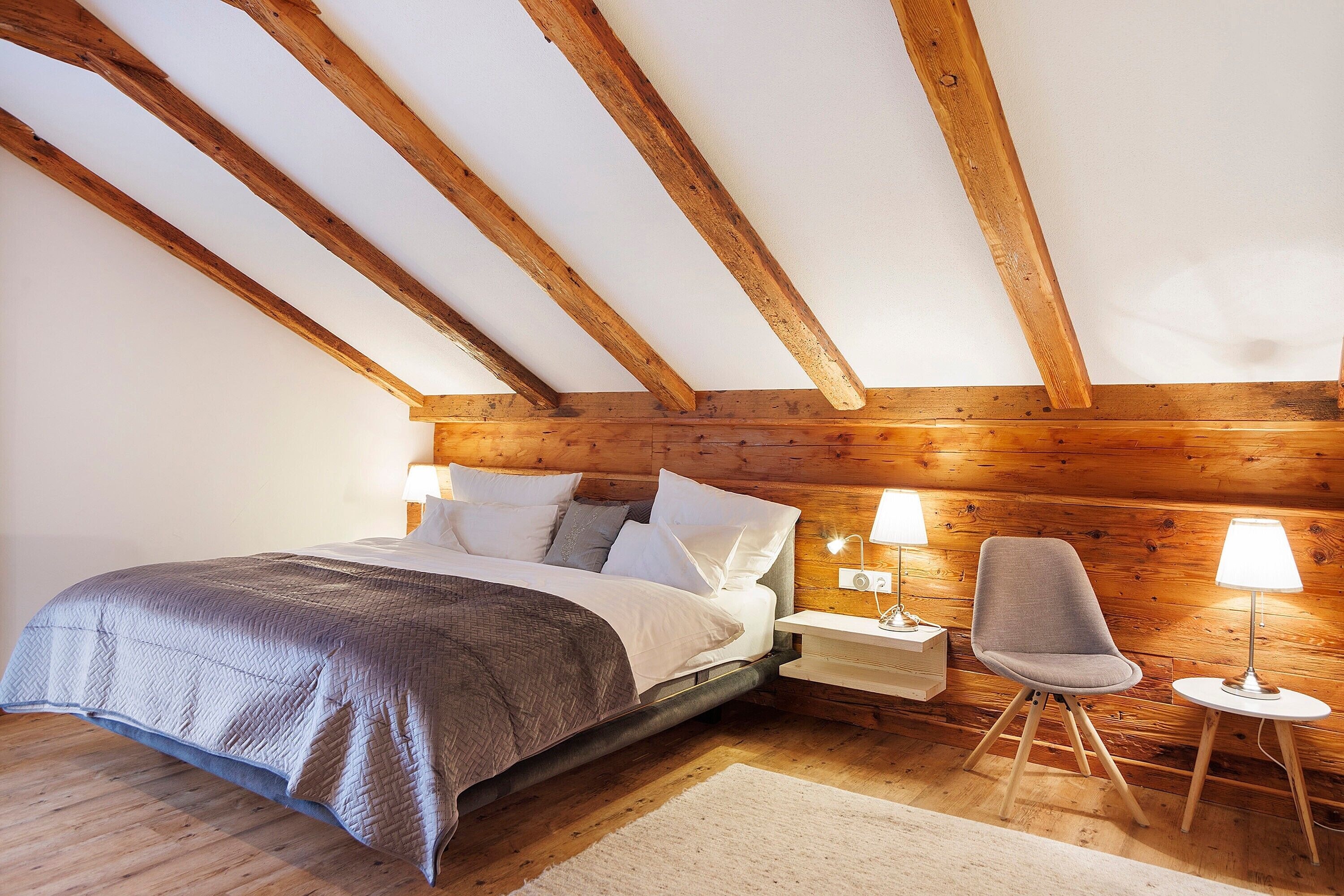 A bedroom with a wooden ceiling, a bed with white bedding, a chair, and two bedside lamps.
