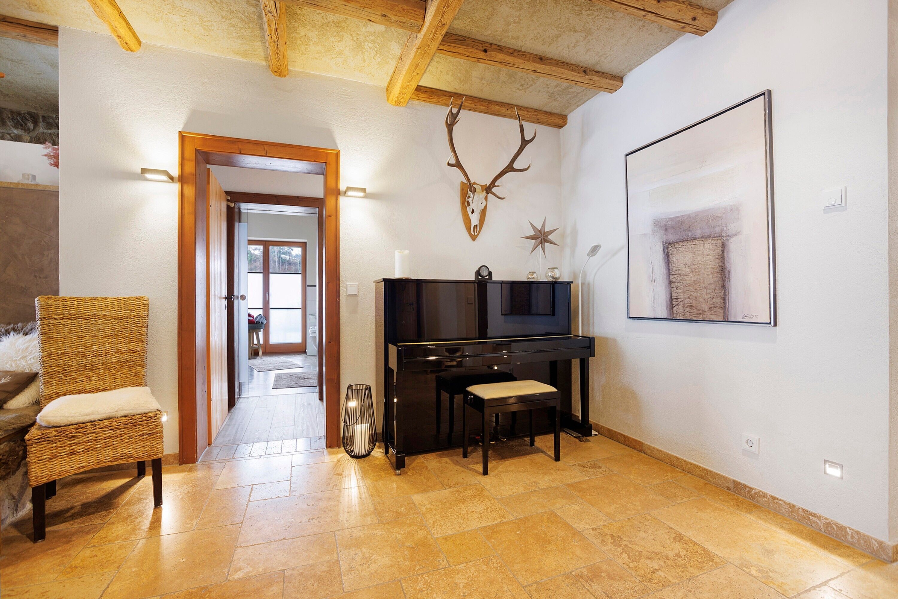 Une pièce comprenant un piano, une chaise, une tête de cerf accrochée au mur, un tableau encadré et une porte donnant sur une autre pièce.