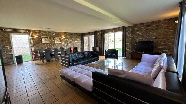 A spacious living area with a large sectional sofa, a glass coffee table, and a television. The room features a brick wall and large windows. - Bungalowpark Landsrade - Vrijstaande bungalow 10 (Gulpen)