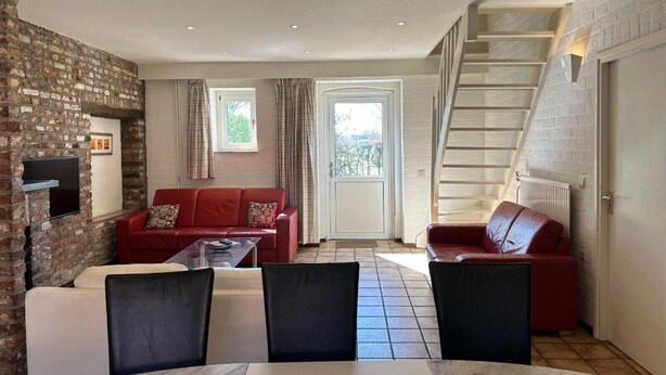 A cozy living room with a red sofa, a coffee table, and a TV mounted on a brick wall. A staircase leads to an upper level. - Bungalowpark Landsrade - Geschakelde Bungalow 9 (Gulpen)