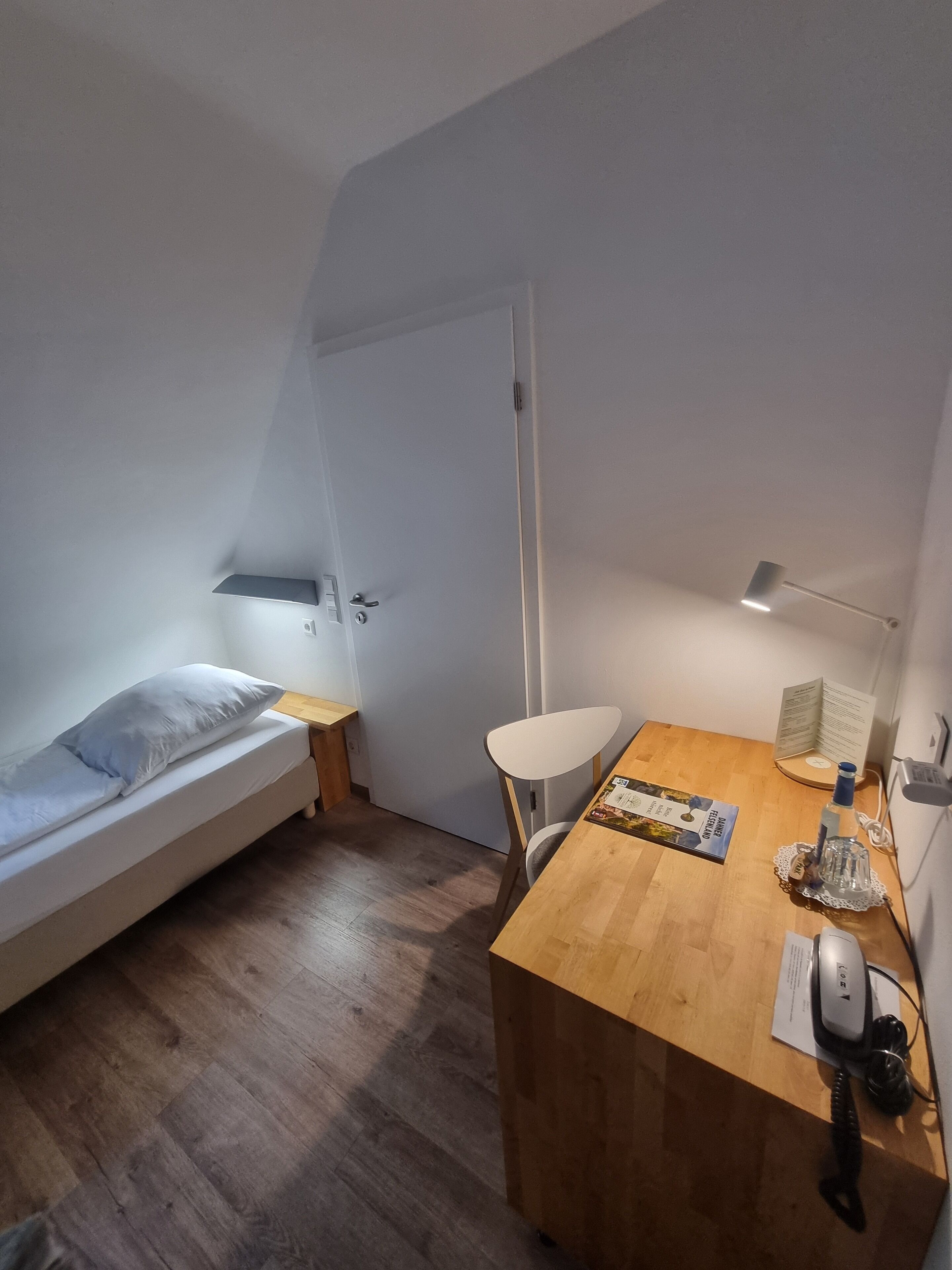 A small, well-lit room with a single bed, a wooden desk, a chair, and a desk lamp.