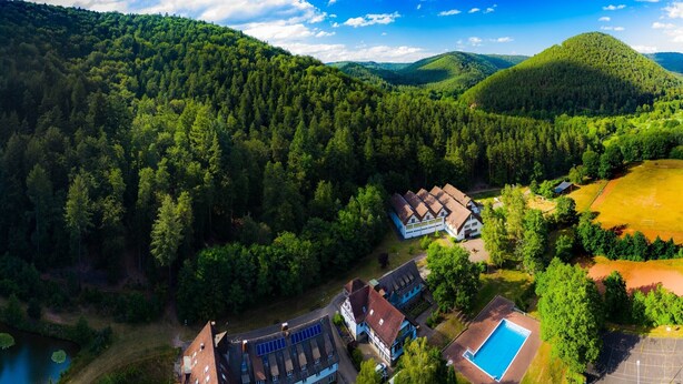 An aerial view of a hotel surrounded by dense forest, with a swimming pool and tennis court. - Die Heilsbach (Schönau)