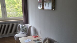 A modern living room with a sofa, armchair, and a small table with a bottle and papers. A window with curtains is visible, and a framed picture hangs on the wall. - Die Heilsbach (Schönau)